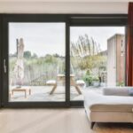 Przez duże okna widać taras z roślinami i drewnianym stołem.