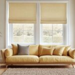 W jasnym salonie znajduje się żółta sofa, a na oknach wiszą beżowe rolety materiałowe.